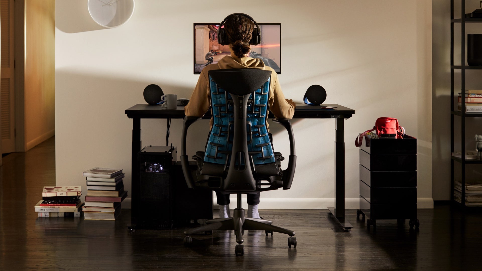 A person sits in the Embody Gaming Chair and looks at a monitor attached to the Ollin Monitor Arm, which sits on top of the gaming desk in a residential setting.